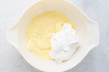 A white mixing bowl containing a smooth yellow batter with a dollop of fluffy cool whip frosting on top, ready to be mixed together.