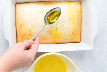 A hand holding a spoon drizzles olive oil over a freshly baked rectangular poke cake in a white baking dish. Below, a white bowl full of olive oil sits on a light gray surface. The cake has small holes poked in it to help absorb the oil.