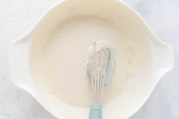 A large white mixing bowl containing batter, with a whisk resting in the mixture. The batter appears smooth and creamy, suggesting it is being mixed or prepared for baking a delicious poke cake. The background is a light, textured surface.