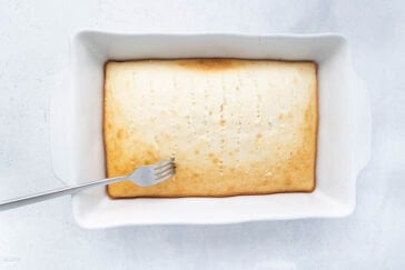 A rectangular baking dish contains a golden-brown, freshly baked poke cake with a fork resting on top. The cake has multiple fork pricks to allow for better absorption of syrup or icing, indicating it's ready for the next step in the recipe.