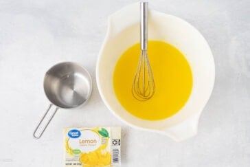 A white mixing bowl containing a yellow liquid and a whisk. Next to the bowl is a stainless steel measuring cup and a Lemon Gelatin Dessert box from the brand Great Value, placed on a light-colored surface.