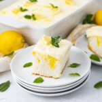 A slice of lemon poke cake topped with creamy icing and garnished with mint leaves, served on a stack of white plates. A whole lemon and the cake in a baking dish are in the background, along with a few scattered mint leaves.