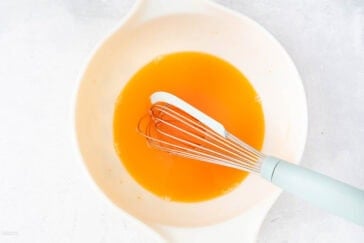 A light-colored mixing bowl containing a whisk and an orange liquid mixture. The whisk with a light blue handle rests partially submerged in the liquid, which appears to be smooth and uniform in texture. The background is a neutral surface.