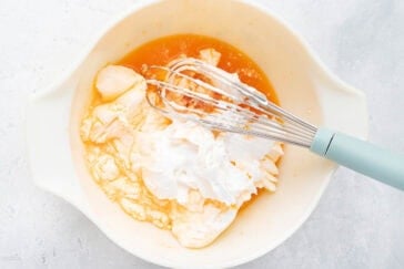 A mixing bowl on a white surface contains a blend of ingredients including sour cream, an orange liquid, and more white ingredients. A whisk with a light blue handle rests in the bowl, partially covered with the mixture.