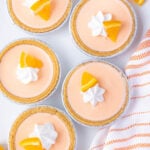 A top-down view of five mini orange cream pies in aluminum tins, each topped with whipped cream and an orange slice. The pies are placed on a white surface next to an orange and white striped cloth.