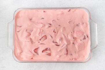 A glass baking dish filled with pink creamy dessert featuring visible slices of strawberries on the surface. The dessert is smooth and spread evenly in the dish, with some strawberry pieces partially submerged, creating a textured appearance.