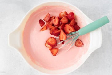 A white mixing bowl containing pink batter and several freshly cut strawberries. A teal whisk is partially submerged in the mixture, resting against the edge of the bowl. The bowl is placed on a light-colored surface.