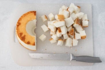 A partially eaten circular sponge cake with a golden brown crust is on a white cutting board. Next to the cake are cubes of the same cake, and a serrated knife with a gray handle rests in front of them. The background is a light gray surface.