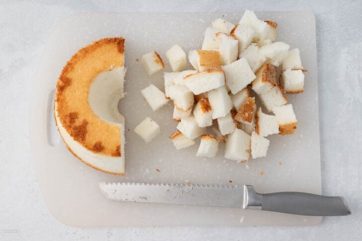 A partially eaten circular sponge cake with a golden brown crust is on a white cutting board. Next to the cake are cubes of the same cake, and a serrated knife with a gray handle rests in front of them. The background is a light gray surface.