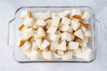A glass baking dish filled with numerous small cubes of white bread, some with golden-brown crusts. The bread cubes are evenly spread out, ready for baking or further preparation. The background is a light, textured surface.