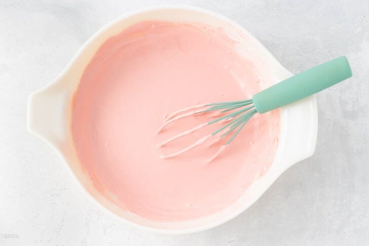 A white mixing bowl filled with smooth, pink batter. A green whisk, slightly covered in batter, rests inside the bowl. The bowl is set against a light, textured background.
