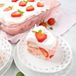A slice of strawberry cheesecake lush with layers of creamy pink filling, fresh strawberry pieces, and whipped topping, garnished with strawberry slices and mint leaves. It is served on a decorative white plate with the remaining dessert in a glass dish in the background.