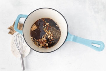 A light blue saucepan with a white interior sits on a white surface. Inside the pan are mixed ingredients including chopped onions, red pepper flakes, ground black pepper, brown sugar, and liquid ingredients. Beside the pan is a small whisk and a wooden trivet.