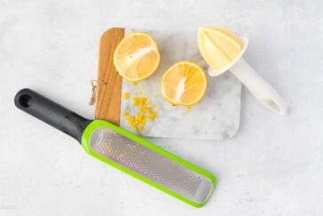 A marble cutting board holds a halved lemon and a lemon reamer with zest scattered around. Near the board, a green and black zester rests on a light gray surface, sprinkled with lemon zest flakes. A wooden handle peeks out from the back of the board.