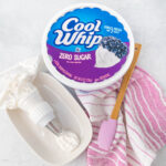 An open tub of Cool Whip Zero Sugar has a small amount of whipped cream scooped out and placed in a piping bag on a white oval dish. A wooden spoon with a pink spatula end rests on a pink and white towel nearby. The background is a light, textured surface.
