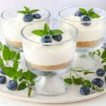 Glass cups filled with creamy no-bake cheesecake, each topped with fresh blueberries and mint leaves. The cheesecake has a layer of graham cracker crust at the bottom. The cups are arranged on a white plate, surrounded by mint leaves and scattered blueberries.