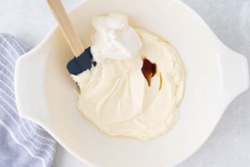A white mixing bowl contains thick, creamy batter with a dollop of cream and a drop of vanilla extract on top. A black silicone spatula with a wooden handle rests in the bowl. A striped gray and white kitchen towel is positioned on the left side of the image.