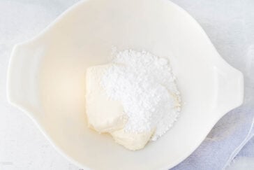 A white mixing bowl containing chunks of cream cheese and a mound of powdered sugar sits on a light surface. Part of a light fabric napkin or cloth is visible to the right of the bowl.