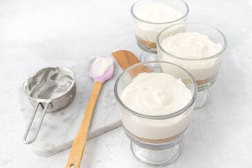 Three glass dishes filled with layered dessert, featuring a crumbly base and creamy topping, are placed on a white surface. Nearby, a marble slab holds a wooden spatula with a pink tip and a metal measuring cup with remnants of the creamy mixture.