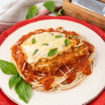 A plate of spaghetti topped with marinara sauce and a browned piece of chicken covered in melted cheese, garnished with fresh basil leaves. The dish is presented on a white plate with a red rim, alongside a cloth napkin and a wooden cheese grater.