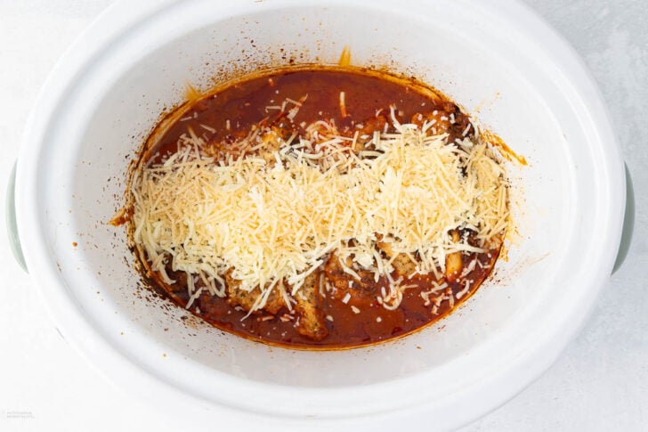 A white slow cooker filled with a rich red tomato-based sauce is topped with shredded cheese. The dish appears partially cooked, with the cheese just beginning to melt into the sauce. The edges of the slow cooker are slightly stained with sauce.