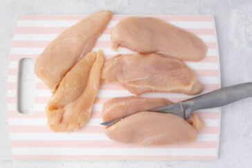 Raw chicken breasts on a striped cutting board with a knife next to them. The cutting board has a handle on the left side and rests on a light-colored surface.