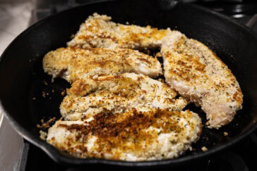 Breaded chicken fillets are frying in a black cast iron skillet. The chicken is golden brown and crispy on the outside, indicating that it is nearly cooked. The skillet sits on a stovetop burner.