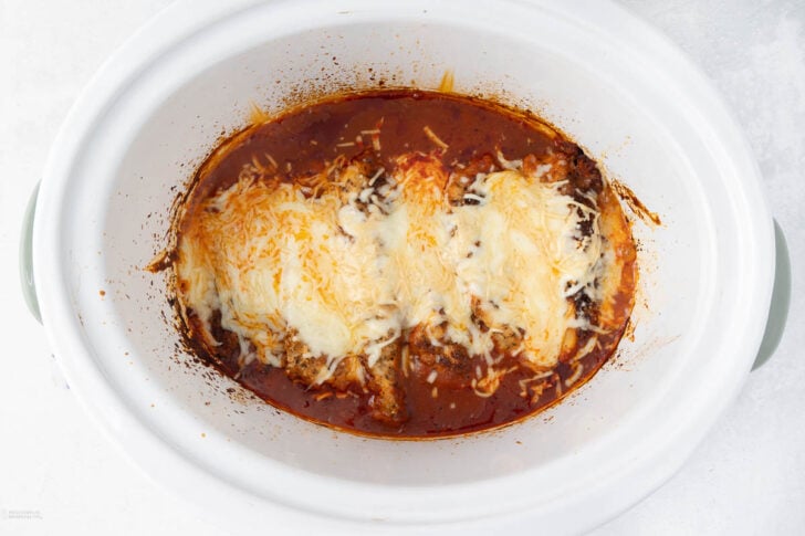 A white oval slow cooker filled with a rich tomato sauce and topped with melted cheese, displaying a baked dish. The edges are slightly browned, suggesting it's hot and freshly cooked. The surface appears to have a mix of cheeses, including mozzarella.