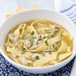 A bowl of chicken noodle soup filled with wide egg noodles, chunks of chicken, and garnished with fresh parsley. The bowl sits on a blue and white patterned cloth, with a hint of crackers visible in the background.