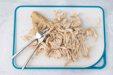 A cooked chicken breast partially shredded with two metal forks sits on a white cutting board with a blue border. The chicken is seasoned, and the shredded pieces are arranged in a pile next to the remaining unshredded portion on the cutting board.