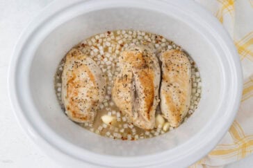 Three cooked chicken breasts seasoned with pepper and spices sit in a slow cooker with diced onions and garlic cloves submerged in broth. The slow cooker is white, and a yellow and white checkered cloth is partially visible on the side.