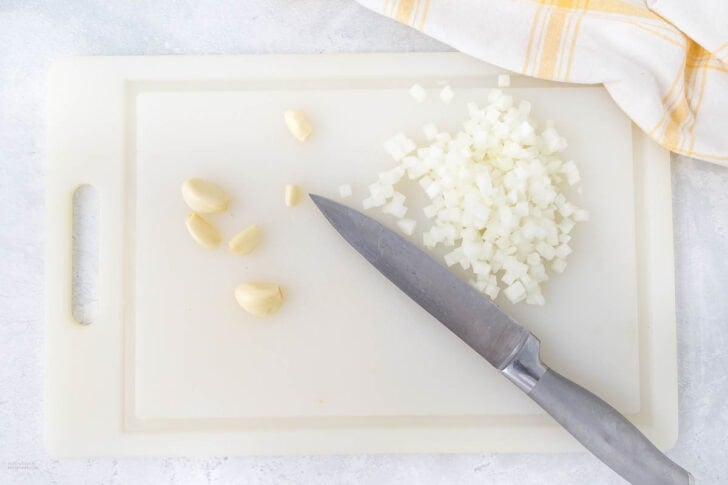 A white cutting board with chopped onions, whole garlic cloves, and a kitchen knife on it—perfect prep for lemon garlic chicken. A yellow and white checkered kitchen towel is partially visible in the corner.