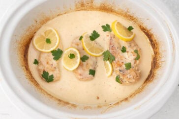 Three pieces of seasoned chicken breast sit in a creamy sauce inside a white baking dish. The chicken is garnished with slices of lemon and fresh parsley leaves. The edges of the dish are slightly browned.