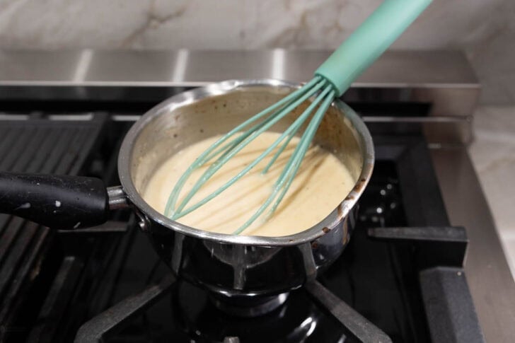 A small stainless steel pot filled with a creamy, light-colored sauce sits on a stove. A mint green whisk is resting inside the pot, partially covered in the sauce, which is being heated. The stove has a black burner and a shiny metallic control panel.