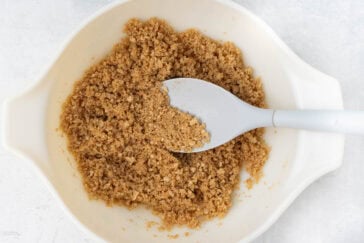 A white bowl filled with light brown sugar is shown from above. A white spoon, slightly coated with brown sugar, rests within the bowl, causing an indentation in the sugar. The background is a light-colored surface.