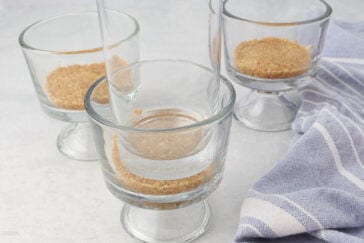 Three glass dessert cups partially filled with a crumbly crust mixture are arranged on a light-colored surface, with a blue and white striped cloth beside them. One cup is positioned sideways, while the other two stand upright.