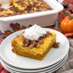 A slice of pumpkin pecan dump cake topped with whipped cream sits on a stack of white plates. The background features a white baking dish with more cake, autumn leaves, and a small decorative pumpkin.