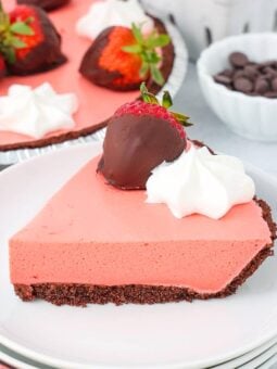 A slice of pink strawberry jello pie with a chocolate cookie crust, topped with a chocolate-covered strawberry and whipped cream, sits on a white plate in front of the whole pie and a bowl of chocolate chips.