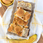 Three slices of banana pudding cheesecake bars are arranged on a rectangular white plate. The bars have a crumbly texture and are topped with round vanilla wafers. A ripe, unpeeled banana sits nearby on a white and yellow checkered cloth.