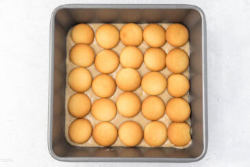 A square baking pan filled with a layer of pudding topped with evenly spaced vanilla wafer cookies. The cookies form a grid pattern, covering the entire surface of the pudding. The background is a light, textured surface.