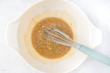 A white mixing bowl contains a brownish mixture being whisked by a manual whisk with a light blue handle. The mixture has a slightly frothy texture, indicating it has been stirred recently. The bowl is placed on a light, textured surface.