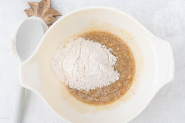 A mixing bowl with dry ingredients, including flour, has been added to a wet mixture. A white spatula with a wooden handle rests to the side of the bowl on a light-colored surface.