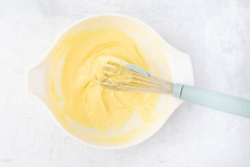 A white bowl filled with creamy yellow cake batter sits on a light grey surface. A whisk with a light blue handle rests inside the bowl, partially coated in the batter. The mixture appears smooth and ready for baking.