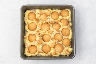 A square baking dish filled with a layered dessert featuring whole vanilla wafers on top of a creamy, pale yellow custard or pudding. Some vanilla wafers are broken into pieces and scattered on the custard. The background is a light-colored surface.