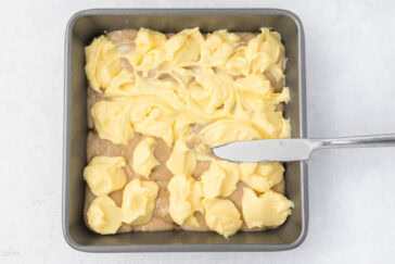 A square baking pan filled with a marbled batter. Dollops of yellow batter are swirled into a lighter, beige batter. A butter knife is partially immersed in the batter, used to create the marbled effect. The pan is placed on a white, textured surface.