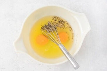 A white mixing bowl contains two cracked eggs, some ground black pepper, and a metal whisk. The bowl is placed on a light, textured surface.