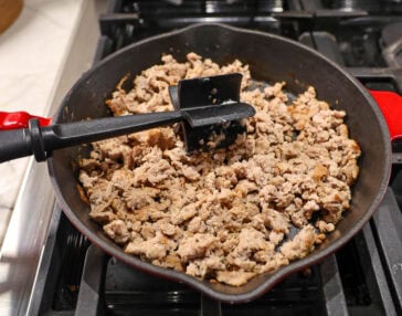 A black skillet on a stovetop contains cooked ground meat. A black spatula rests in the skillet, positioned at an angle. The skillet has red handles, and the stovetop burners are visible underneath.