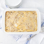 A white rectangular casserole dish filled with egg casserole, featuring a speckled, golden-brown top, sits on a white surface. A blue and white striped towel is partially underneath, and a stack of small white plates is nearby.