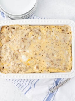 A white rectangular casserole dish filled with egg casserole, featuring a speckled, golden-brown top, sits on a white surface. A blue and white striped towel is partially underneath, and a stack of small white plates is nearby.