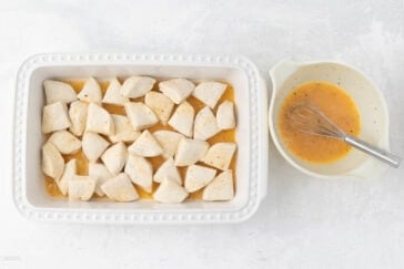 A rectangular white baking dish filled with uncooked biscuit dough pieces sits to the left of a white mixing bowl containing a glaze mixture, and a whisk rests inside the bowl. The background is a light, textured surface.